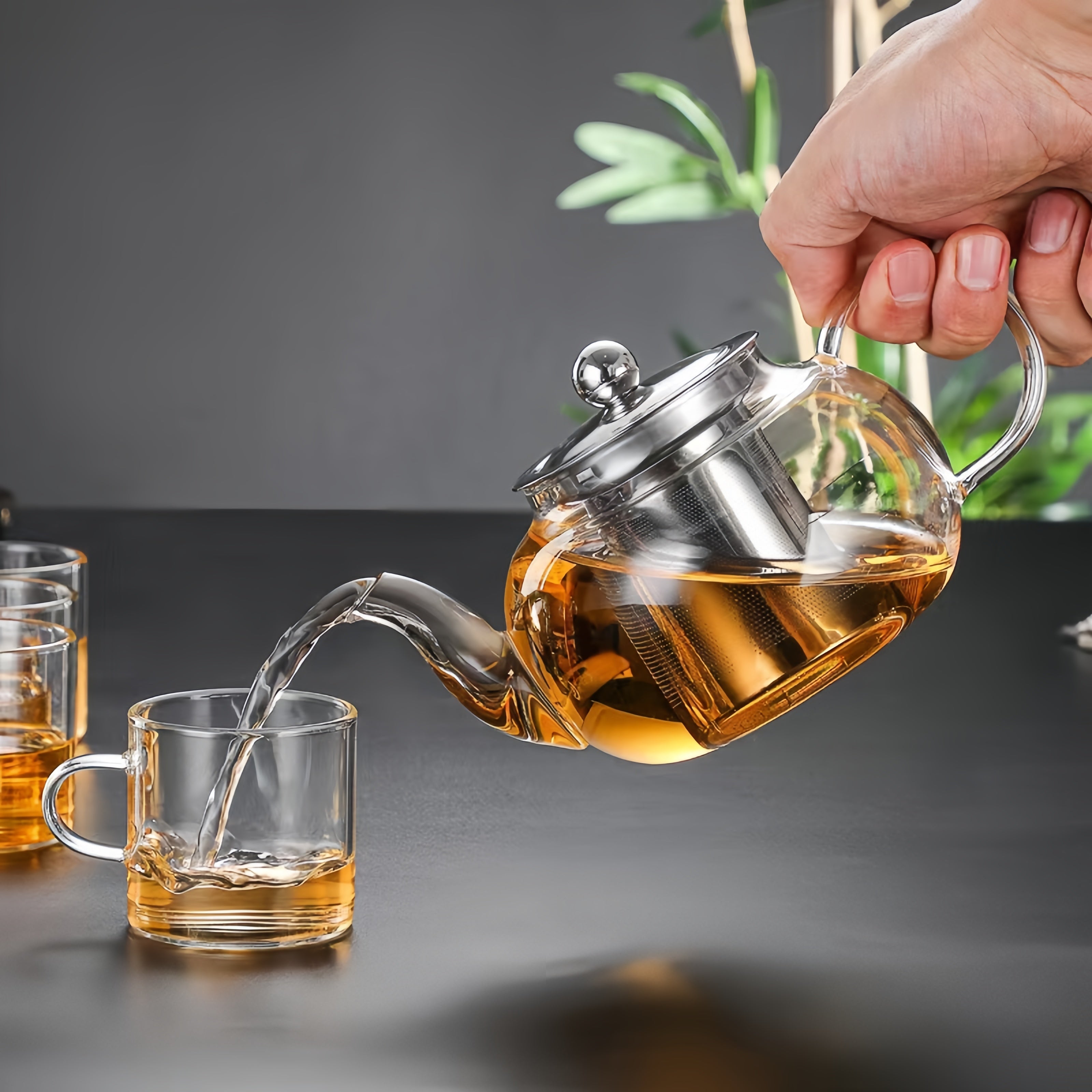 A person pouring tea from a clear glass teapot with a stainless steel lid into a glass cup, with another glass cup beside it.