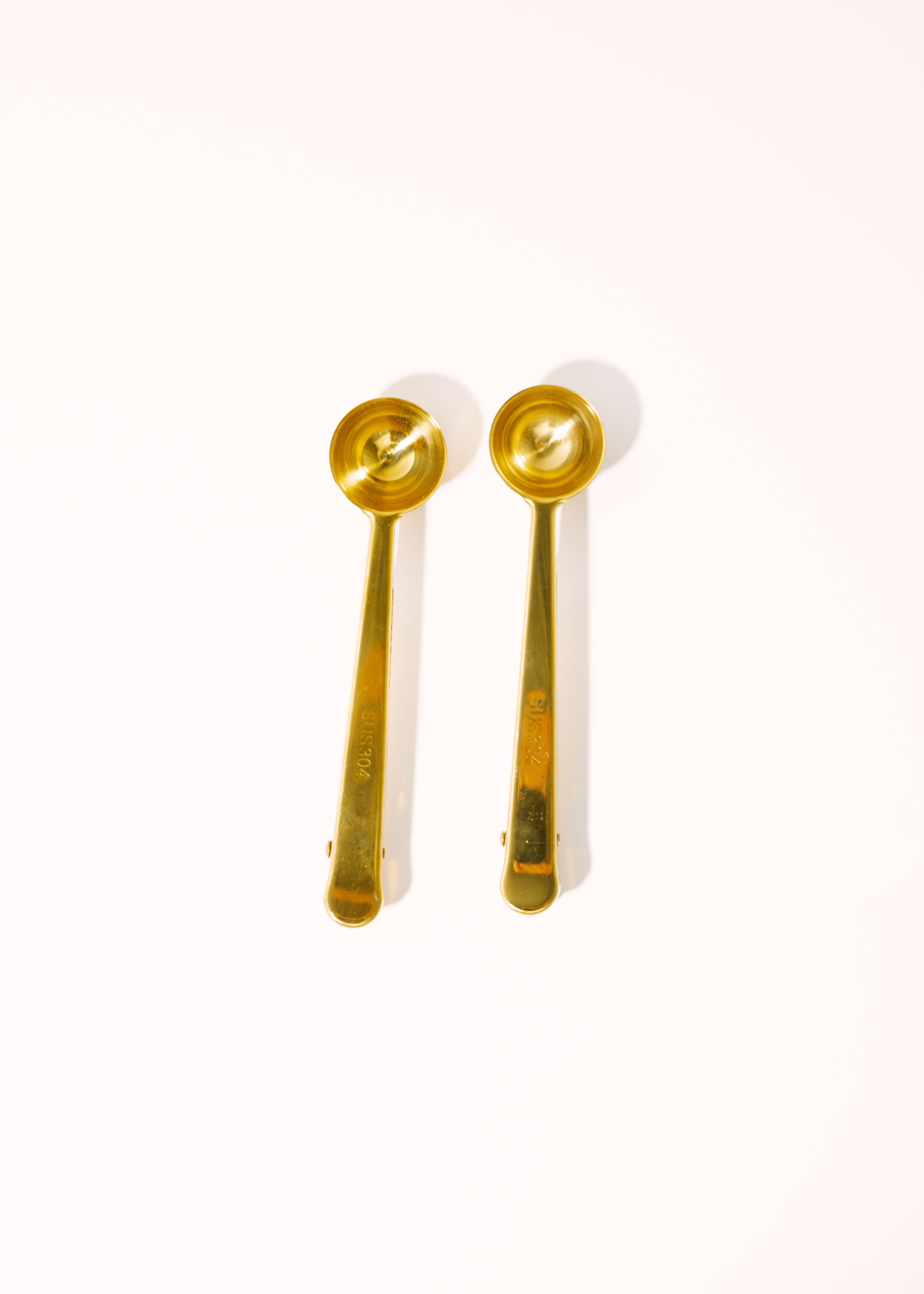 A pair of gold-colored metal measuring spoons with long handles and circular bowls, shown on a plain white background.