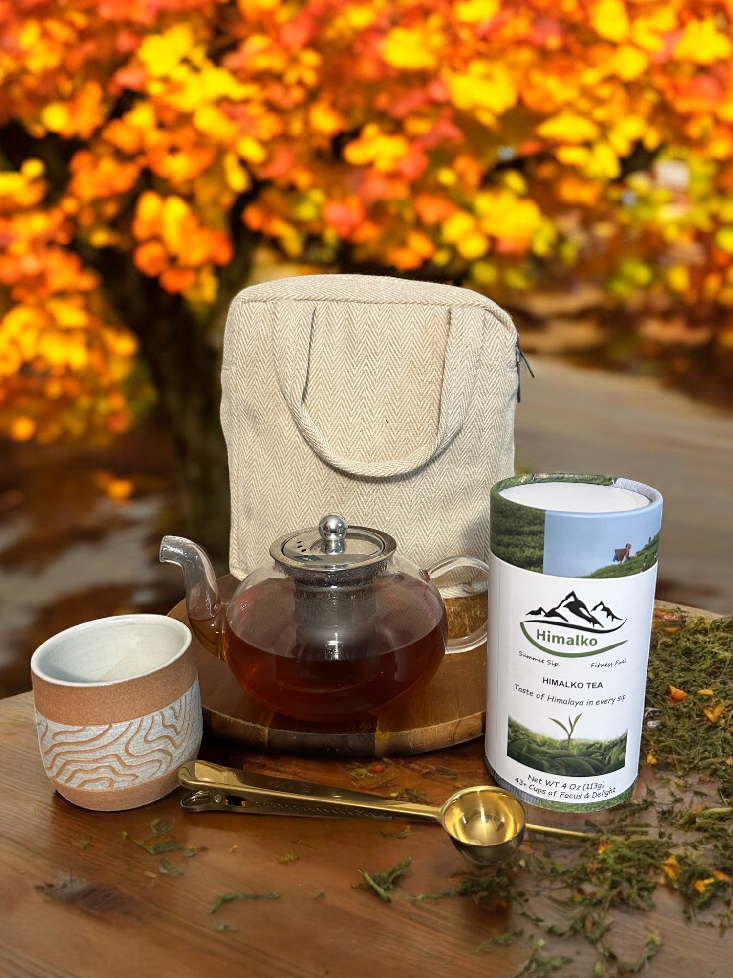 A tea gift set displayed outdoors, including a clear glass teapot with a removal infuser, a handcrafted stoneware teacup, a tea scoop, a gift bag, and a container of Himalko ICED tea.