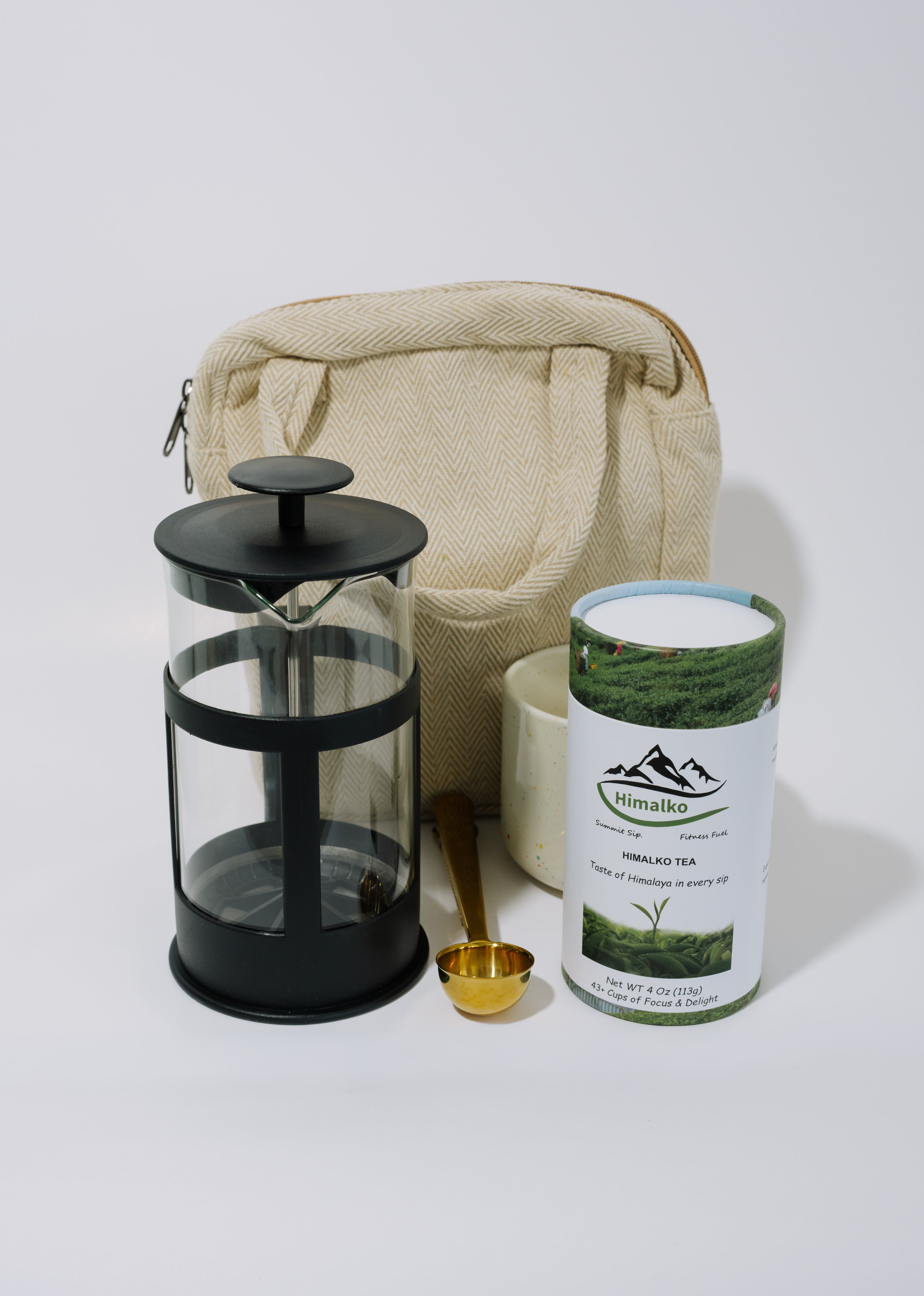 A tea brewing set on a light gray background, including a black glass and metal French press, a beige canvas bag, a white ceramic mug, a gold spoon, and a canister of Hillsdale Organic Green Tea.