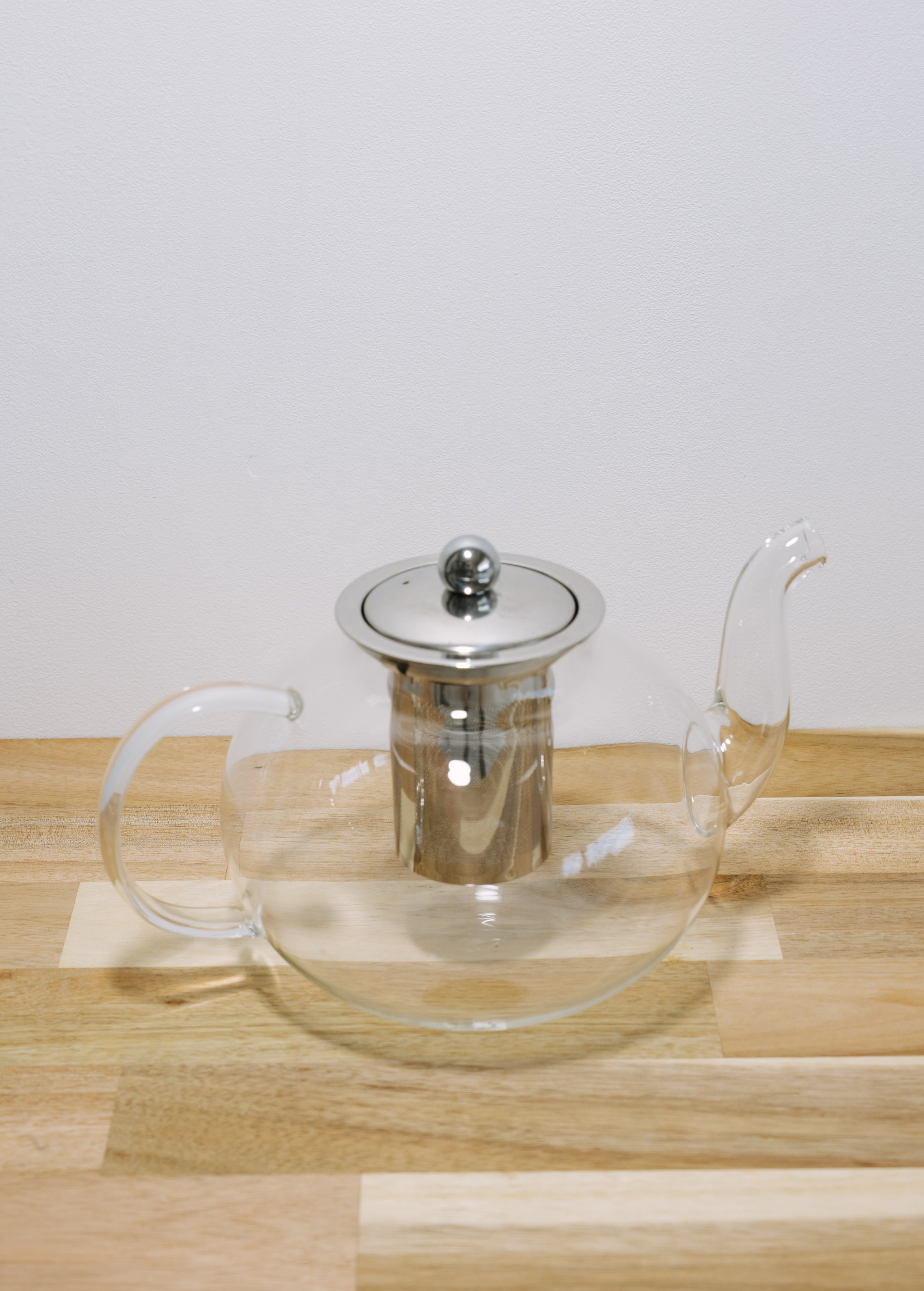 A clear glass teapot with a silver metal lid and infuser, resting on a light wood surface against a plain white background.