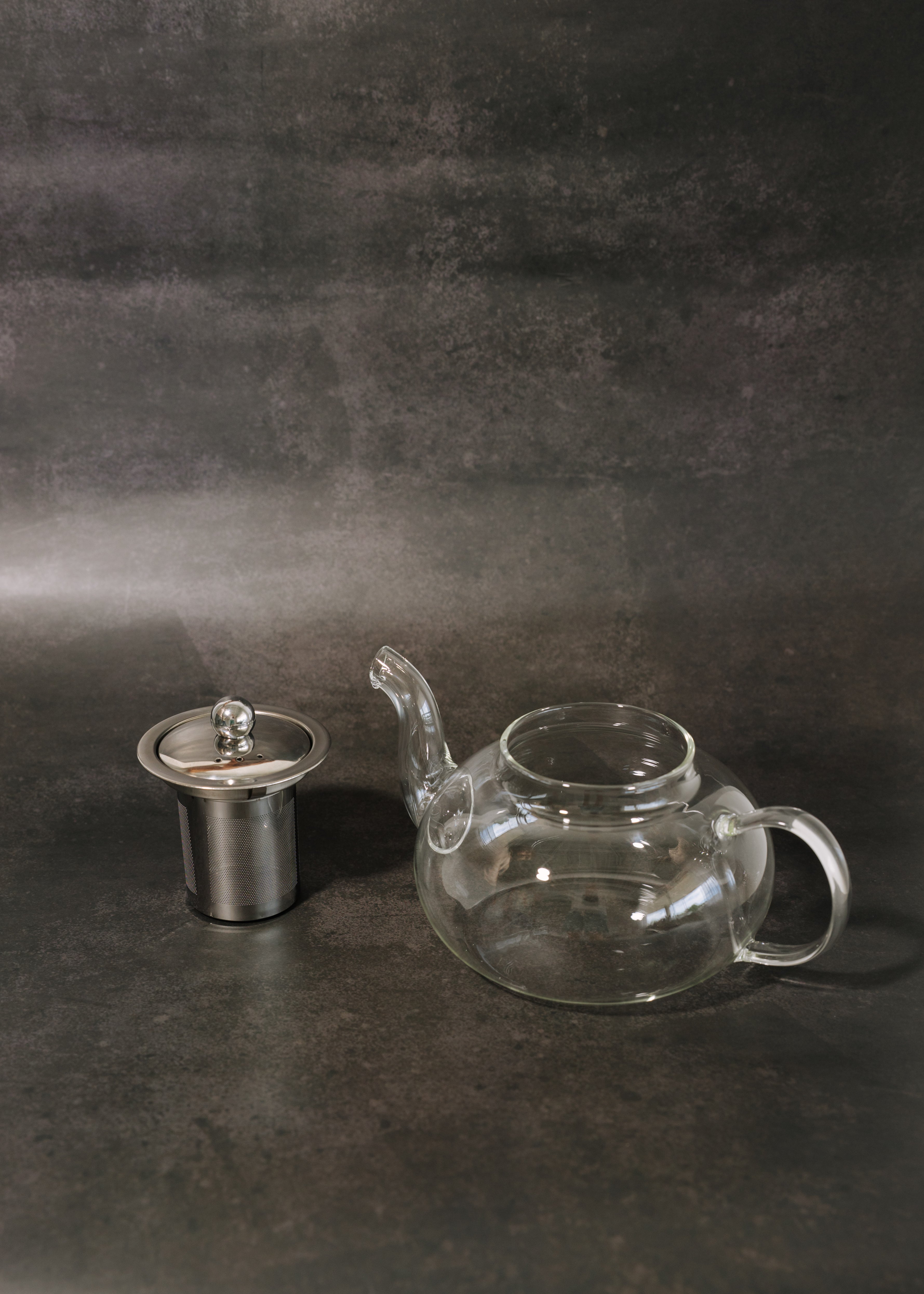 Clear glass teapot with a matching glass lid and a stainless steel infuser filter, displayed on a dark textured background.