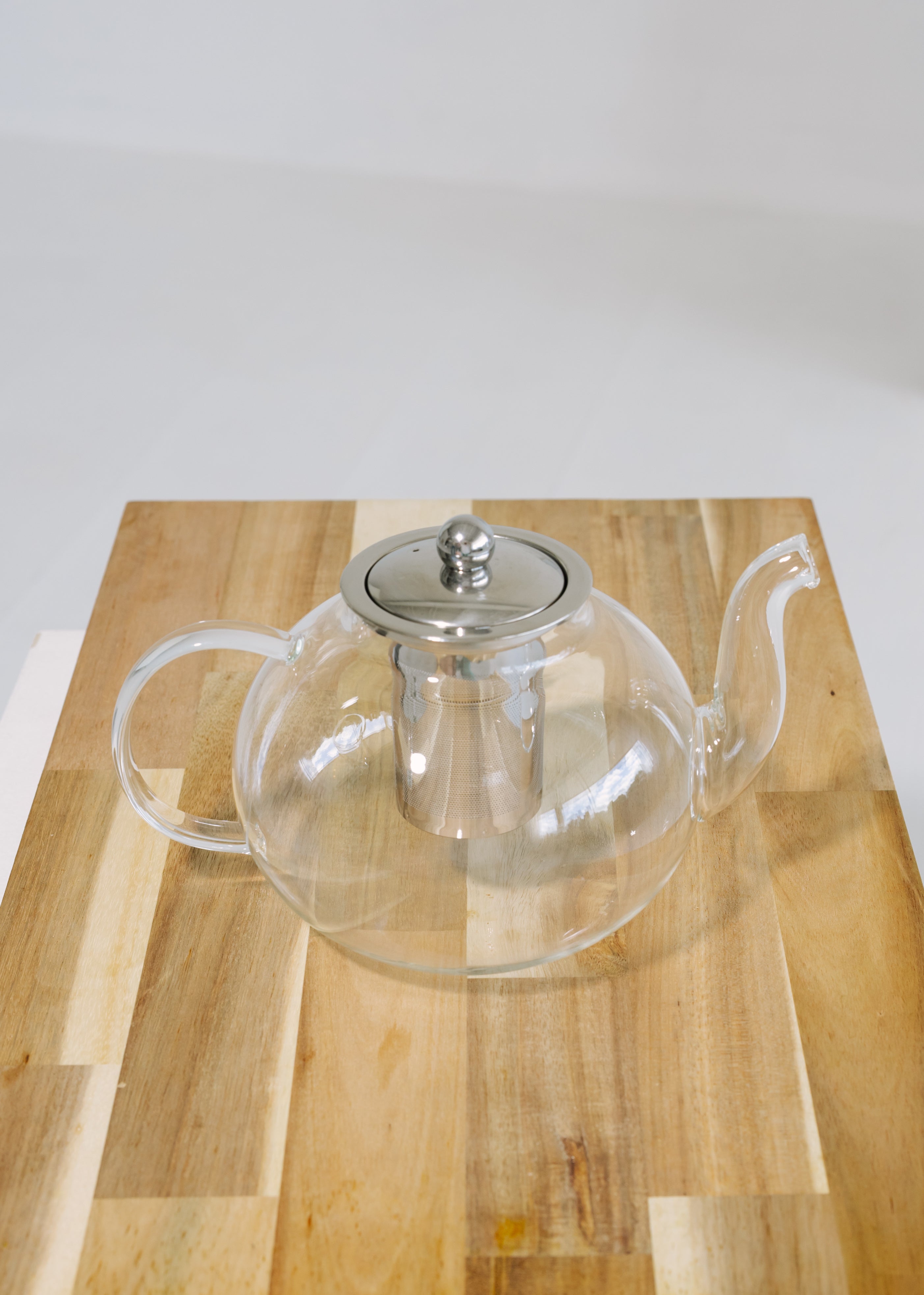 Clear glass teapot with a stainless steel lid and infuser, resting on a wooden cutting board against a white background.