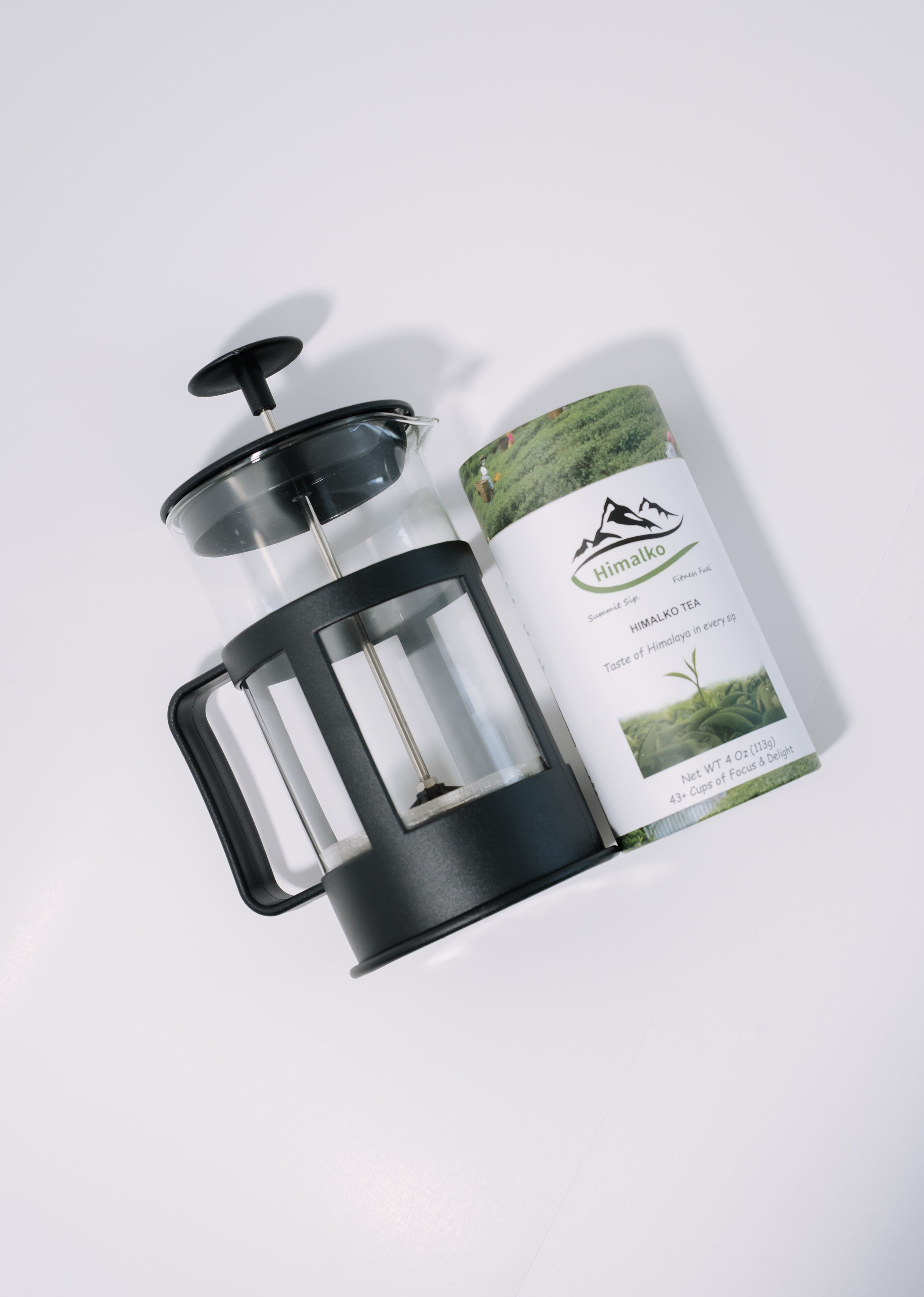 A black French press with a clear glass chamber is shown next to a cylindrical tin of Mountain Organic Herbal Tea on a clean white background.