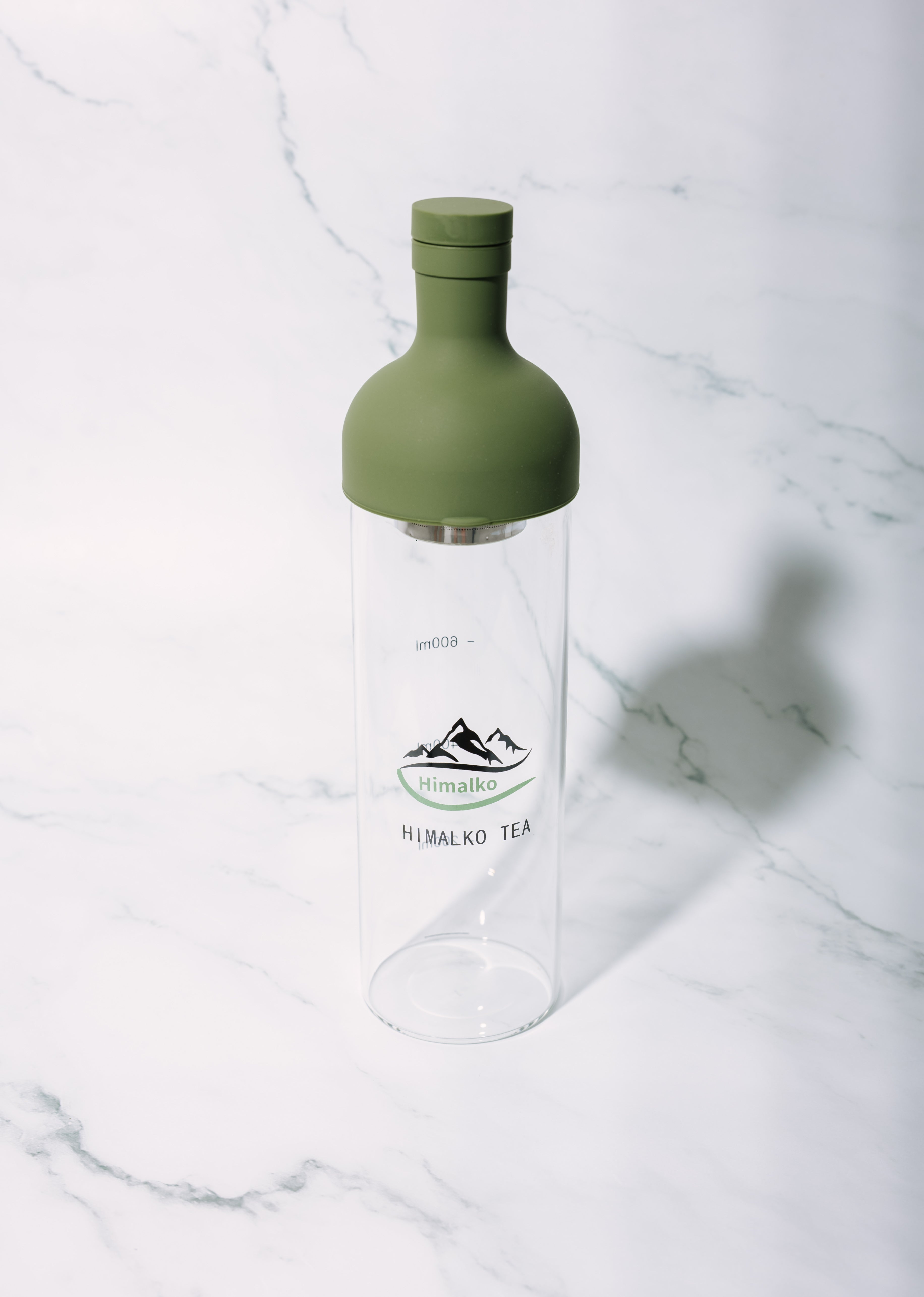 A clear glass bottle with an olive green silicone cap, featuring the 'Himalko' brand logo printed on the front, set on a white marble surface.