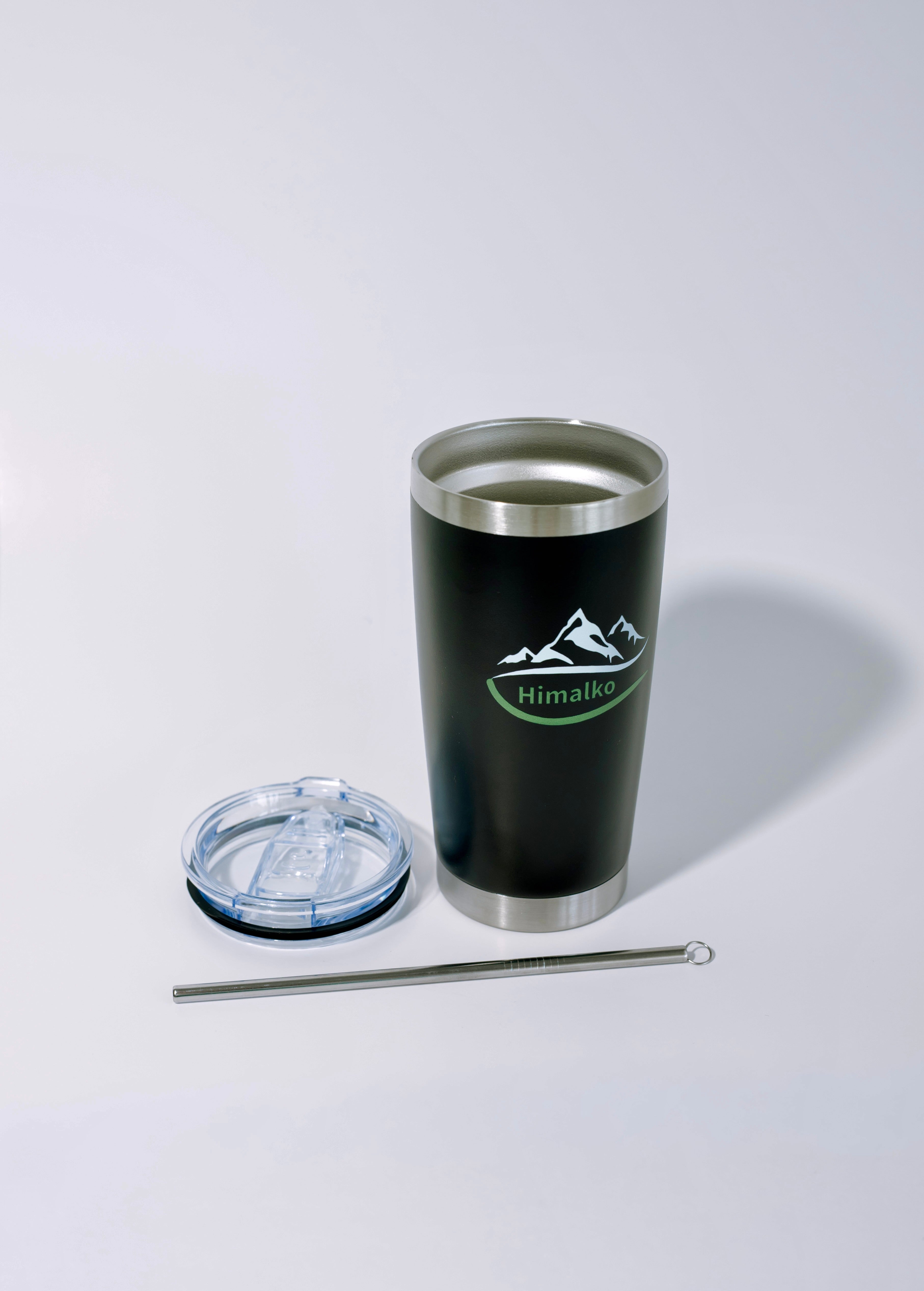 A black insulated travel tumbler with a stainless steel rim and base, featuring a green and white mountain logo with the text 'Himalayas'. It is shown with a clear lid and a metal straw on a light gray background.