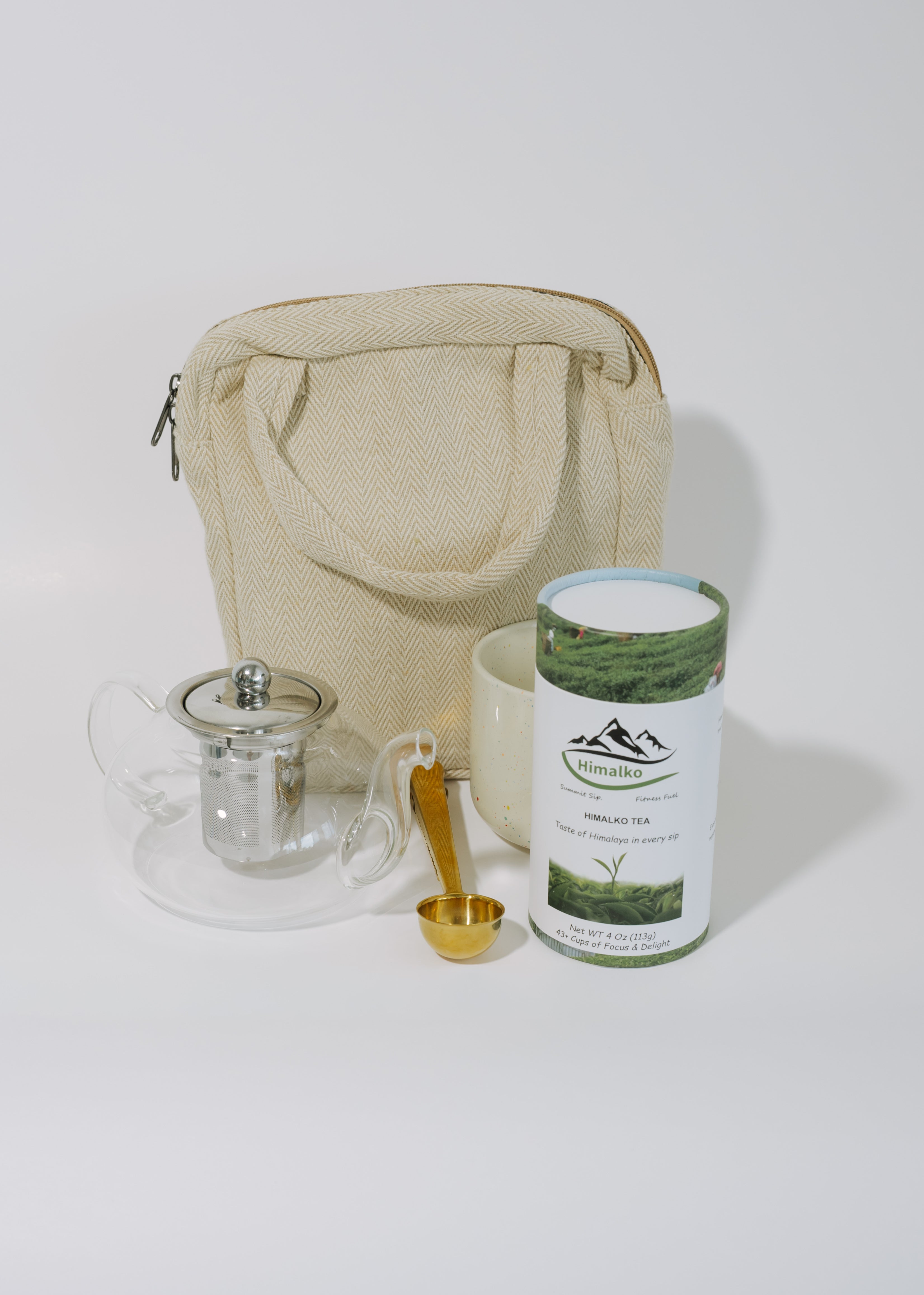 A tea brewing set arranged on a light gray studio background, including a beige canvas tote bag, a clear glass teapot with metal infuser, a white ceramic mug, a small gold spoon, and a cylindrical tin of tea with a green-and-white label.