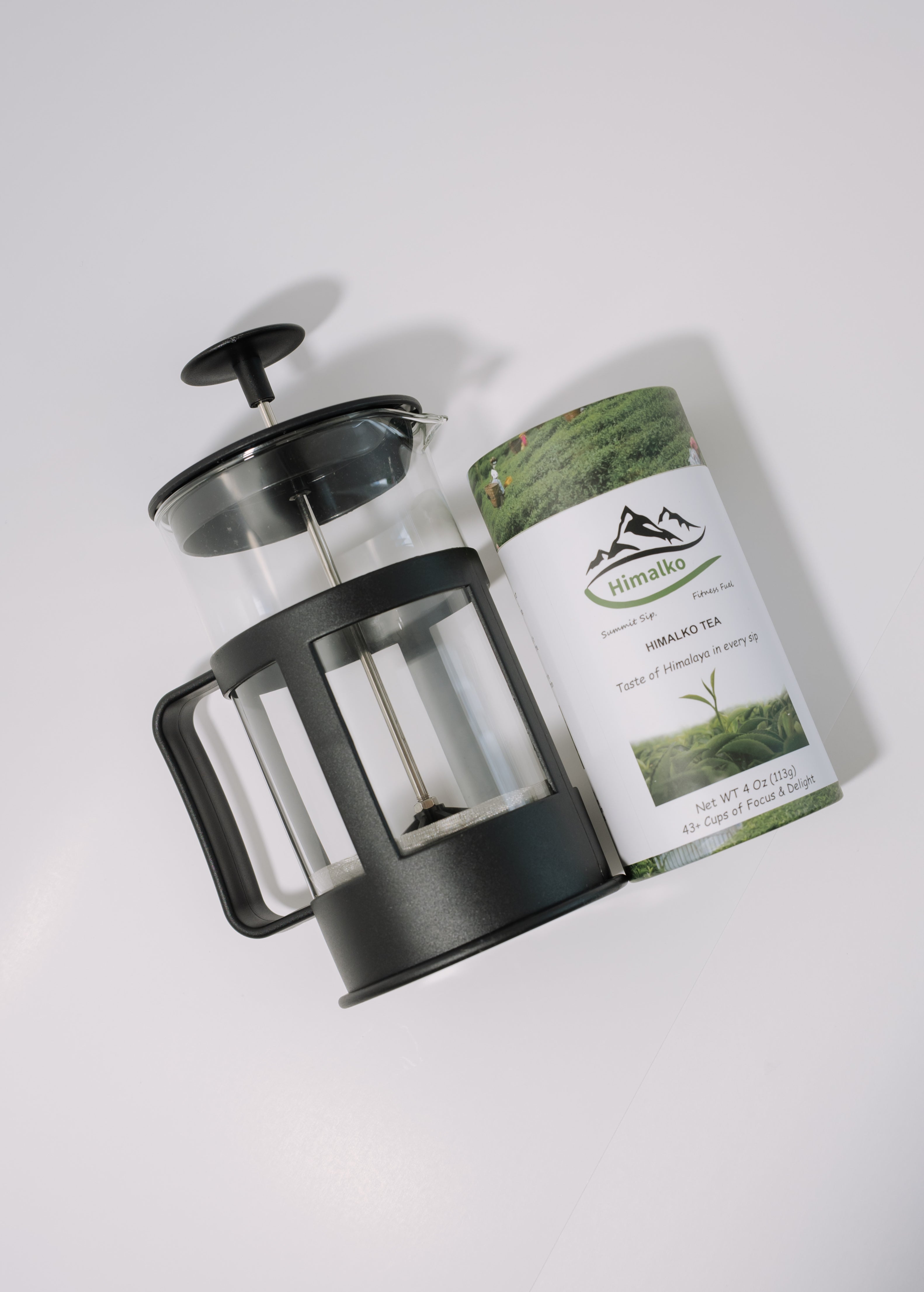 A glass and black French press tea maker positioned next to a cylindrical paper tea canister labeled 'himalaya organic whole leaf tea' on a clean white background.