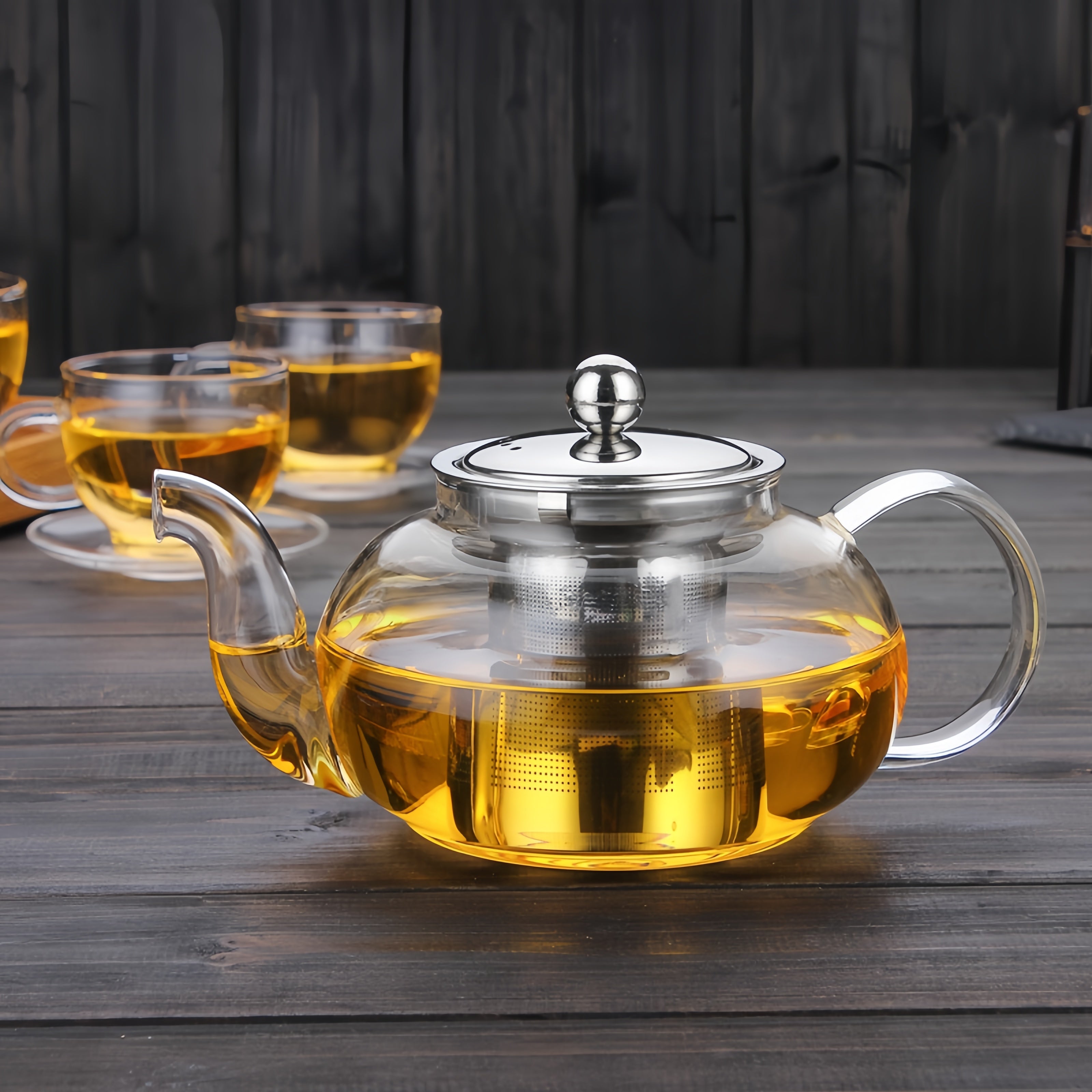 Clear glass teapot with silver lid and handle on a wooden surface, with cups of tea in the background.