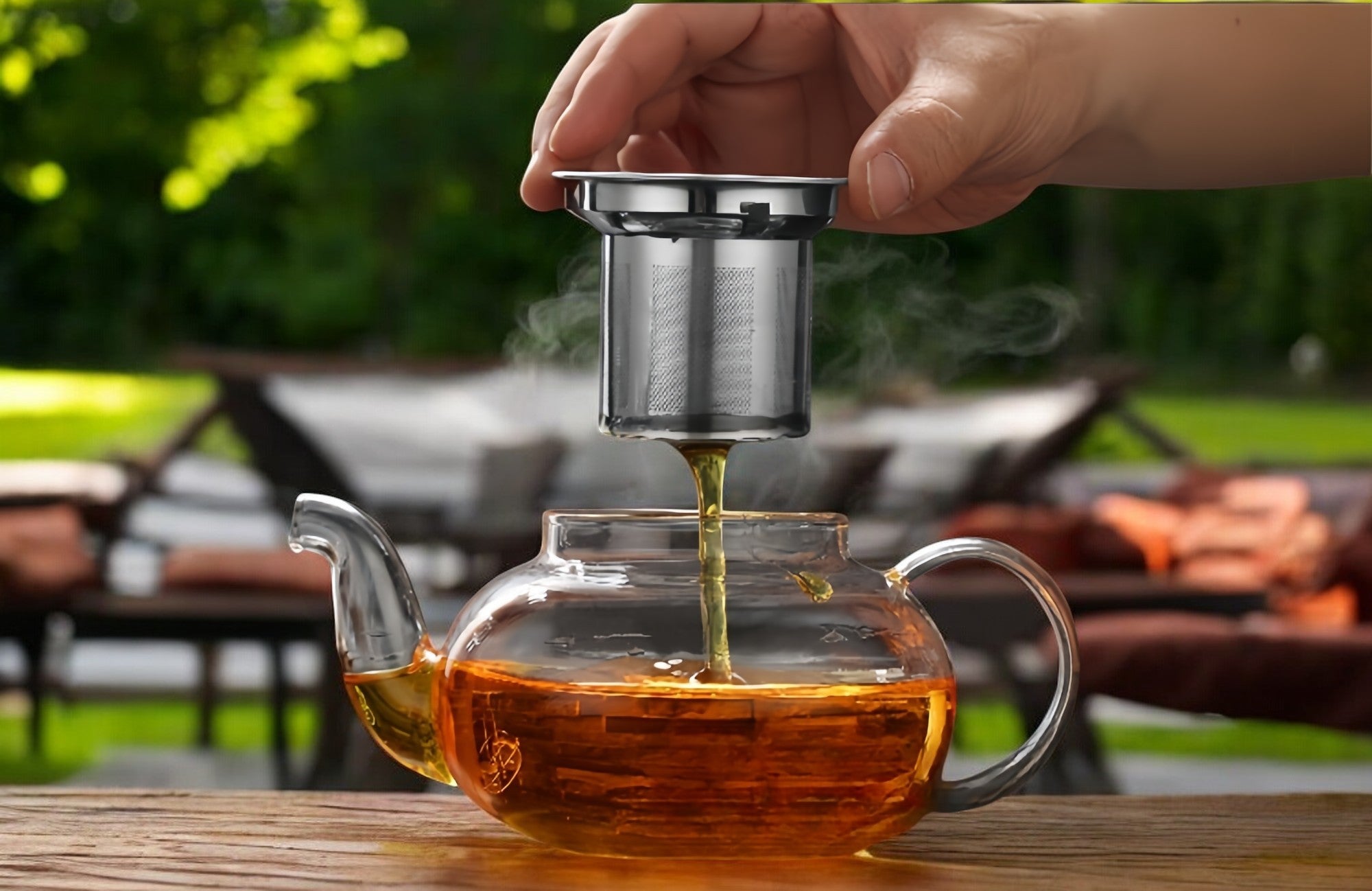 Tea being poured from a glass teapot into a metal tea infuser held by a hand outdoors.
