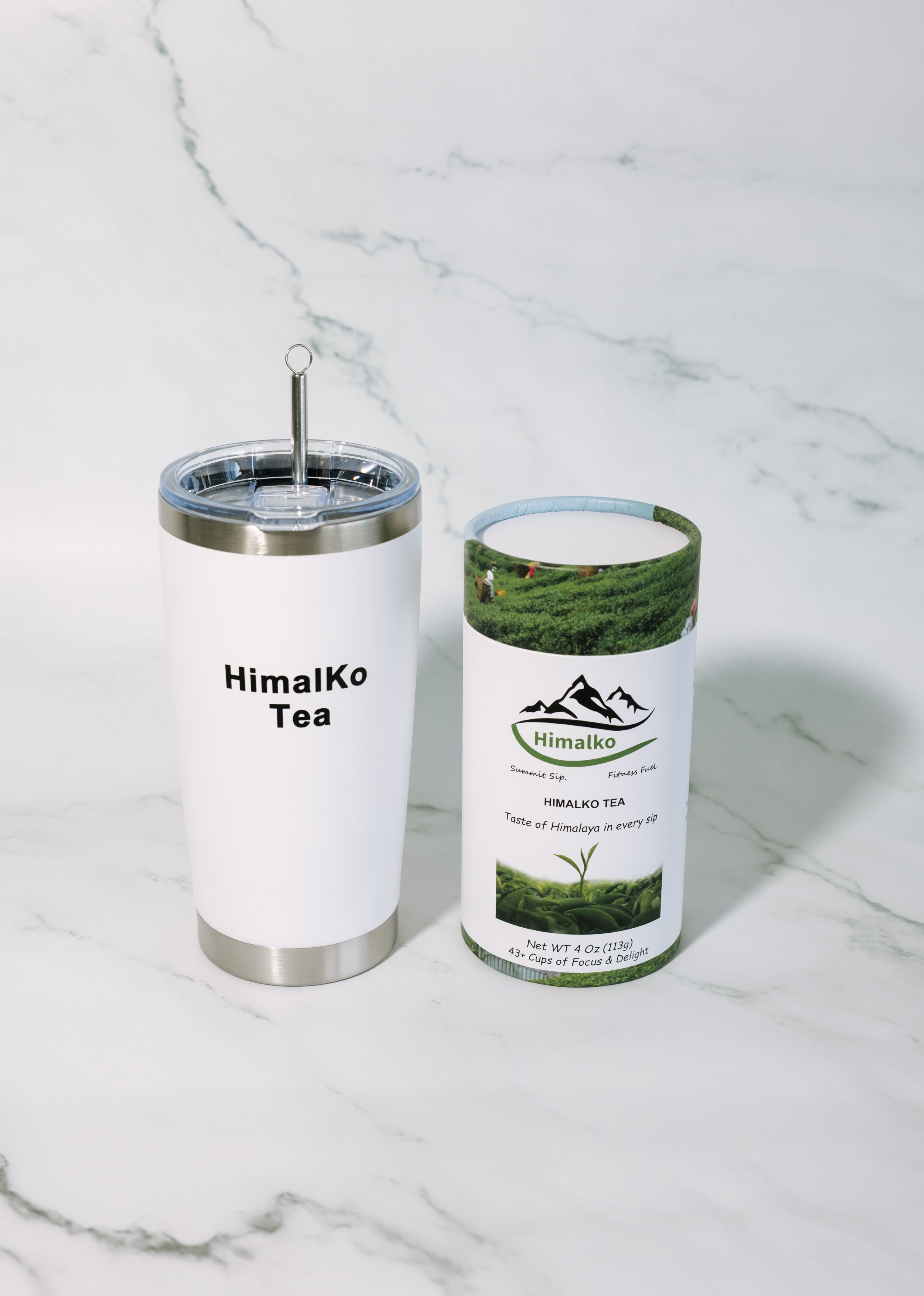 A white stainless steel tumbler with a clear lid and metal straw stands beside a cylindrical canister of HimalKo Tea on a white marble surface.